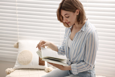 Woman reading book near window blinds at home