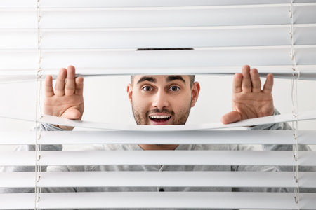 Young man looking through window blinds on white background