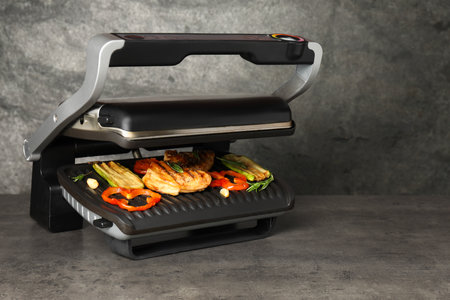Electric grill with tasty meat, spices and vegetables on gray textured table, space for textの写真素材