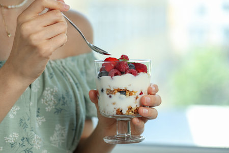 Woman eating tasty yogurt with fresh berries and granola indoors, closeup. Space for textの写真素材