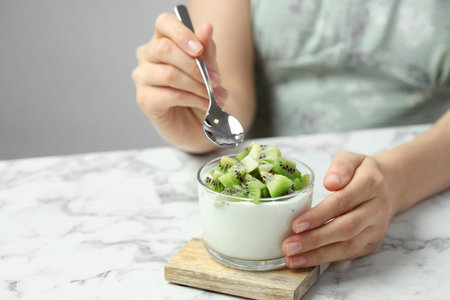 Woman eating tasty yogurt with fresh kiwi at white marble table, closeup. Space for textの写真素材