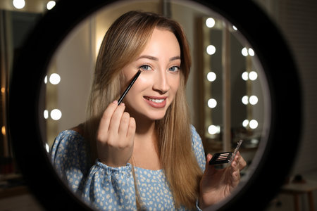 Beautiful young woman applying eye shadow with brush indoors, view through ring lamp
