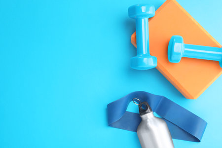 Two dumbbells, yoga block, fitness elastic band and thermo bottle on light blue background, flat...