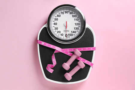 Scale tied with measuring tape and dumbbells on pink background, top view