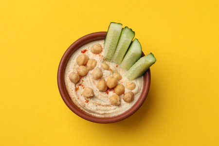 Tasty hummus with chickpeas and cucumber in bowl on yellow table, top viewの写真素材