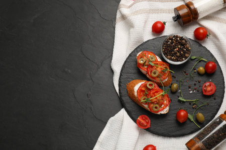 Delicious ricotta bruschettas with sliced tomatoes, olives and greens on black table, top view....