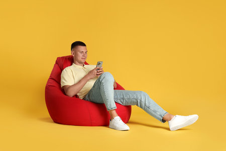Handsome man with smartphone on red bean bag chair against yellow backgroundの写真素材