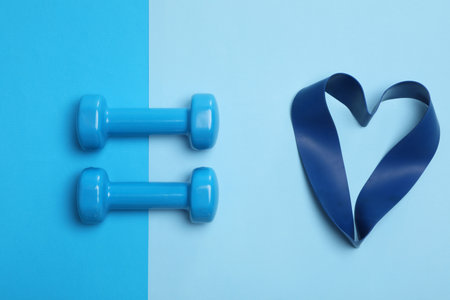 Two dumbbells and fitness elastic band in shape of heart on light blue background, flat lay. Lovely workoutの写真素材