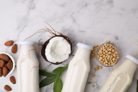 Different types of vegan milk in bottles with ingredients and leaves on white marble table, flat layの写真素材