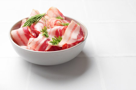 Slices of raw bacon and rosemary in bowl on white tiled table, space for textの写真素材
