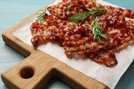 Slices of tasty fried bacon and rosemary on light blue wooden table, closeup