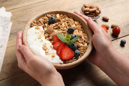 Woman holding bowl of tasty granola with berries, almond and yogurt at wooden table, closeupの写真素材