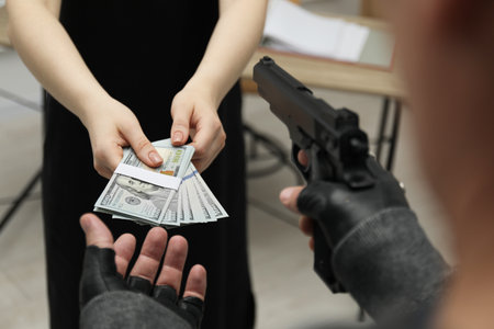 Woman giving money to criminal with gun indoors, closeup. Armed robbery