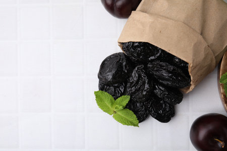 Paper bag with delicious prunes, fresh ripe plums and mint on white tiled table, flat lay. Space for textの写真素材