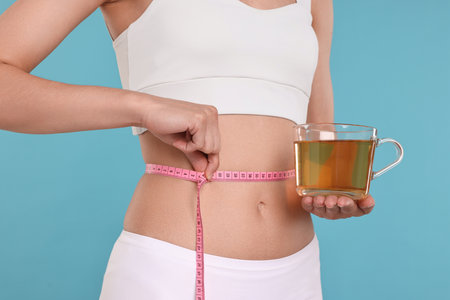 Weight loss concept. Woman with cup of diet tea and measuring tape on light blue background, closeupの写真素材