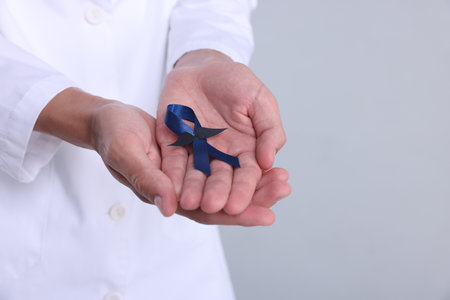 Prostate cancer awareness. Doctor holding blue ribbon with fake mustache on light gray background, closeup. Space for textの写真素材