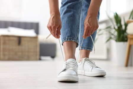 Woman tying shoelace of white sneaker indoors, closeup. Space for text