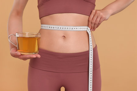 Weight loss concept. Woman with cup of diet tea and measuring tape on beige background, closeupの写真素材