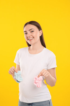 Expecting twins. Pregnant woman holding two pairs of socks on yellow backgroundの写真素材