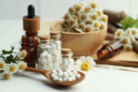 Homeopathic remedy. Spoon with pills, bottles and chamomile flowers on white table, closeupの写真素材