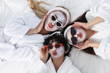 Happy friends with facial masks and sunglasses on bed, top view. Spa partyの写真素材