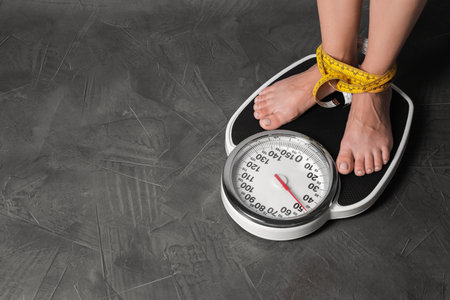 Eating disorder. Woman tied with measuring tape standing on floor scale indoors, closeup. Space for textの写真素材