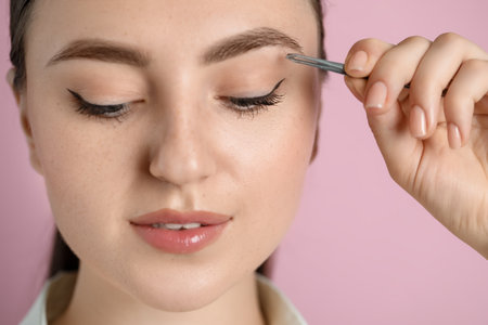 Young woman plucking eyebrow with tweezers on pink background, closeup
