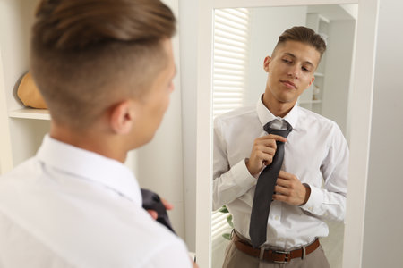 Handsome man adjusting necktie near mirror indoorsの写真素材