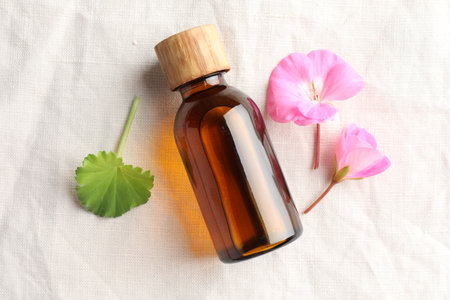 Bottle of geranium essential oil and beautiful flowers on white fabric, flat layの写真素材