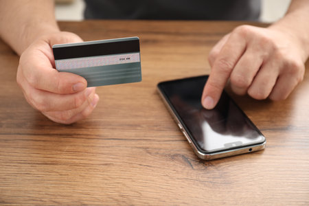 Man with credit card using smartphone at wooden table indoors, closeupの写真素材