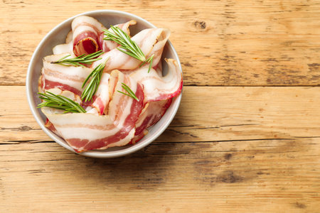 Slices of raw bacon and rosemary in bowl on wooden table, top view. Space for text