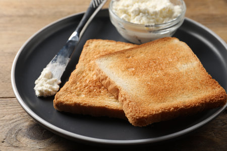 Delicious toasted bread slices with cream cheese and knife on wooden table, closeup