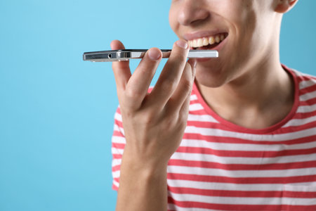 Young man recording voice message via smartphone on light blue background, closeup. Space for text
