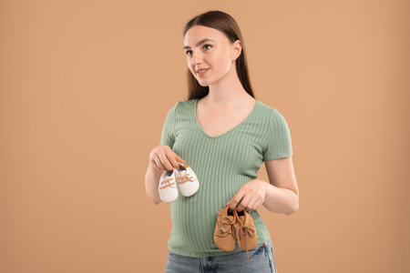 Expecting twins. Pregnant woman holding two pairs of shoes on light brown background