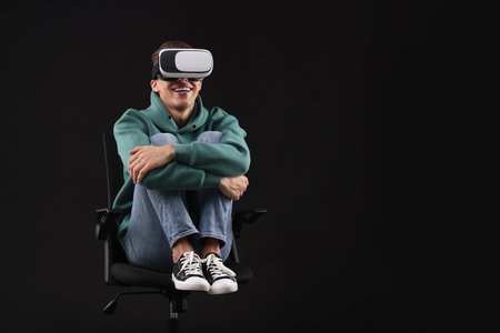 Happy young man with virtual reality headset sitting on chair against black background, space for...