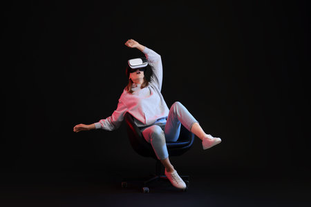 Emotional woman with virtual reality headset sitting on chair in neon lights against black...