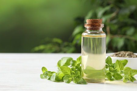 Essential oil in bottle and oregano leaves on white wooden table against blurred green background, closeup. Space for textの写真素材