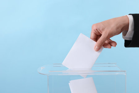 Woman putting her vote into ballot box against light blue background, closeup. Space for textの写真素材