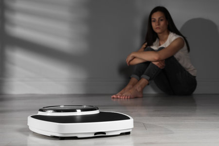 Eating disorder. Sad woman sitting near wall indoors, focus on scaleの写真素材