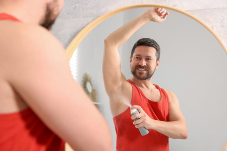 Smiling man applying spray deodorant near mirror at home