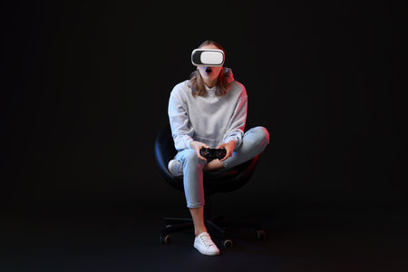 Emotional woman with virtual reality headset and controller sitting on chair in neon lights against black backgroundの写真素材