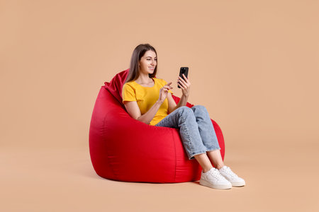 Smiling woman using smartphone while sitting on red bean bag chair against beige backgroundの写真素材
