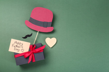 Card with phrase Happy Father's Day, gift box, heart, moustache and paper hat on green background, flat lay. Space for textの写真素材