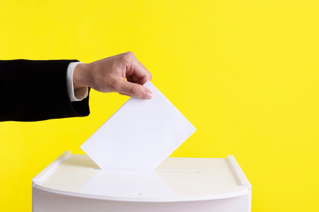 Woman putting her vote into ballot box against yellow background, closeup. Space for text