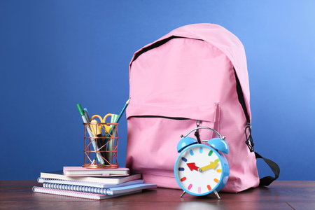 Backpack with different school stationery and alarm clock on wooden table against blue backgroundの写真素材