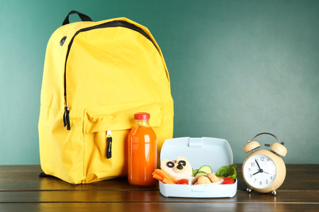 Backpack, lunch box with snacks, bottle of juice and alarm clock on wooden table against color backgroundの写真素材