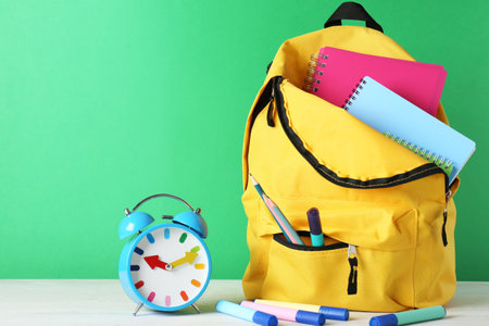 Backpack with different school stationery and alarm clock on white table against green background