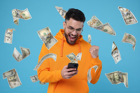 Happy man using mobile phone under money rain on light blue background