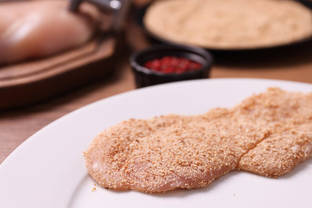 Making schnitzel. Plate with raw slice of meat in bread crumbs on wooden table, closeupの写真素材