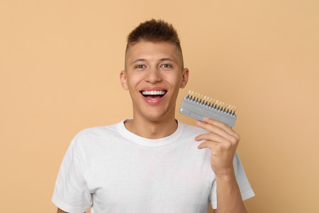 Happy young man with teeth color samples on beige background. Dental veneersの写真素材